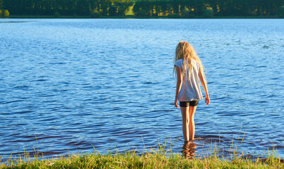 Wall Mural - Alone child blond girl standing in the water in the light of the setting sun in summer