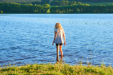 Wall Mural - Alone child blond girl standing in the water in the light of the setting sun in summer