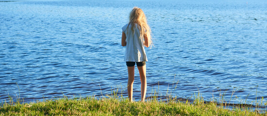 Wall Mural - Alone child blond girl standing in the water in the light of the setting sun in summer