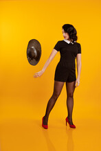 Young Brunette Woman In A Short Black Dress Holds A Black Hat With Large Brim And Poses On A Yellow Background