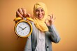 © Krakenimages.com - Middle age brunette business woman wearing muslim traditional hijab holding alarm clock doing ok sign with fingers, excellent symbol
