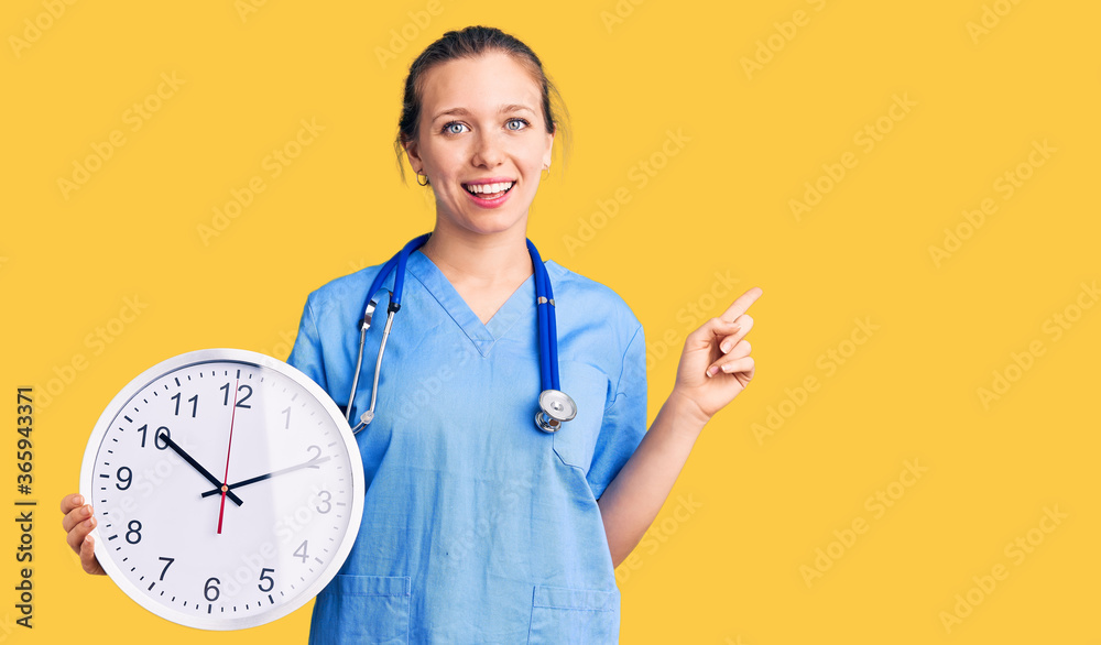 Young beautiful blonde woman wearing doctor uniform and stethoscope smiling happy pointing with hand and finger to the side