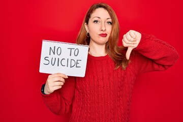 Beautiful redhead woman holding banner showing no to suicide message over red background with angry face, negative sign showing dislike with thumbs down, rejection concept