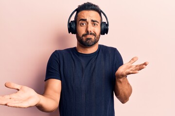 Young hispanic man listening to music using headphones clueless and confused expression with arms and hands raised. doubt concept.