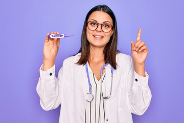 Young beautiful brunette doctor woman wearing stethoscope and coat holding thermometer surprised with an idea or question pointing finger with happy face, number one