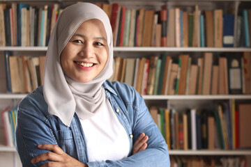 Wall Mural - Portrait of cheerful Asian muslim female librarian wearing hijab and smiling, woman standing against books in library