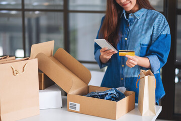 Canvas Print - Closeup image of a beautiful young woman using mobile phone and credit card for online shopping with shopping bag and postal parcel box of clothing on the table