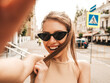 © halayalex - Portrait of beautiful smiling brunette girl in summer clothes. Model taking selfie on smartphone.Woman making photos in warm sunny day in the street in sunglasses. Cheerful female going crazy