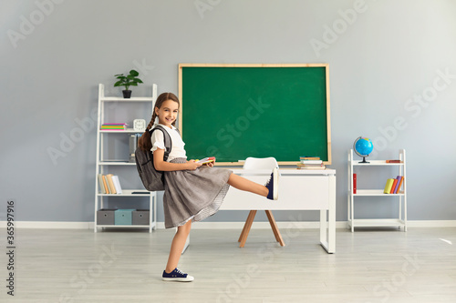 Cute girl in uniform with backpack walking in classroom, copy space text. Elementary pupil starting new school year
