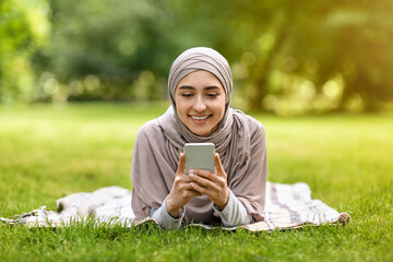 Wall Mural - Pretty girl in hijab laying on grass, using phone