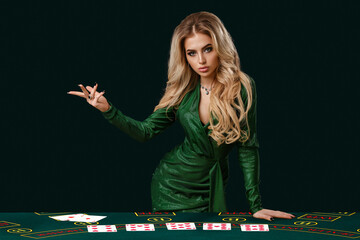 Girl in stylish dress is showing something, leaning on playing table with cards on it, posing on green background. Poker, casino. Close-up, copy space