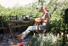 Portrait Of An Elderly Woman Playing  Guitar And Watching  Online Lessons  On Laptop While Practicing In The Garden. Senior Woman Happy And Enjoy Life After Retired. Online Training
