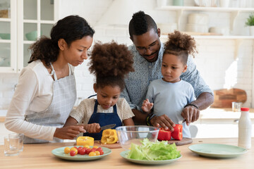 Loving african American mom and dad teach cooking chop vegetables with excited little children in kitchen, happy caring biracial parents prepare food organic salad with small ethnic kids at home