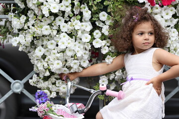 little curly girl with a bicycle on a city street near a flower bed.