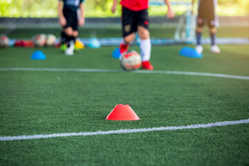 Wall Mural - selective focus to red cone marker is soccer training equipment on green artificial turf with blurry kid players training background. Material for trainning class of football academy.