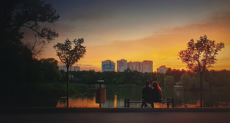 Young couple sitting together on the bench in the park near the lake against bright sunset. Romantic lifestyle background, togetherness concept. Lovers spend time together in a relaxing, calm evening.