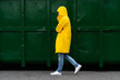 Man in a yellow raincoat walks down the street in the rain weather next to green container, side view. Outdoor. 
