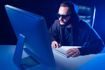 Poster - Hacker in a hood on a dark background. Harker face illuminates light of a computer. Male hacker and computer in a neon light. Cyber criminal is looking at monitor. Computer network hacker.