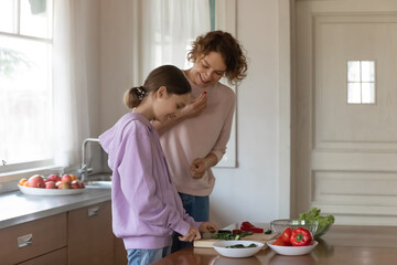 Happy mother and teenager daughter cooking salad, standing in modern kitchen at home together, smiling teen girl cutting fresh vegetables with knife, overjoyed mum and child preparing dinner