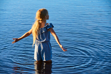 Wall Mural - Alone child blond girl standing in the water in the light of the setting sun in summer