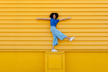 Happy Young Woman Balancing On Platform In Front Of Yellow Wall