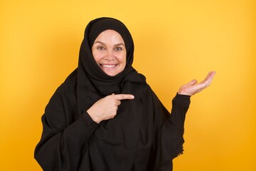 Wall Mural - Beautiful middle aged muslim woman wearing black hijab over yellow background holding hand showing advertisement