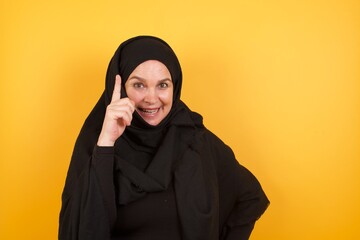 Wall Mural - Beautiful middle aged muslim woman wearing black hijab over yellow background holding finger up having idea and posing