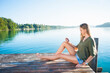 © Westend61 - Young woman sitting on jetty at lake looking at cell phone
