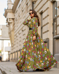 Full length outdoor portrait of young elegant woman wearing green dress with floral design and walking at city street on a summer day