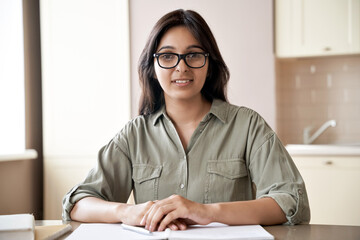 Smiling young indian female teacher wearing glasses sitting at table. Indian woman school tutor or university student studying working from home office giving online remote classes concept. Portrait