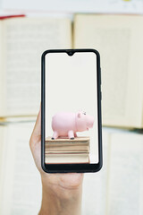Wall Mural - cost of online education. person hold smartphone with image of piggy bank and books. copy space