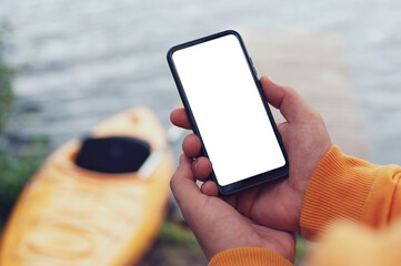 The tourist holds a phone in his hands. Mock up smartphone close-up on the background of a kayak and a lake. Concept on the topic of tourism and recreation.