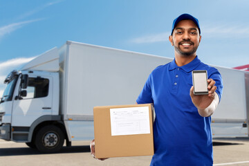 Wall Mural - mail service, technology and shipment concept - happy indian delivery man with smartphone and parcel box in blue uniform over truck on street background