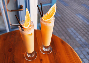 Wall Mural - Two tropical cocktails adorned by fresh orange and brightly lit by setting sun on the wooden table of a seafront mediterranean cafe. Copy space.