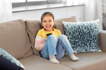 people and childhood concept - happy smiling little girl with smartphone sitting on sofa at home