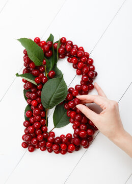 Letter B from summer harvest of cherry on white wooden background. Fruit summer alphabet. Sweet mood font. Woman hand is making a letter B from green leaves and cherry berries. Colorful ABC, Text 