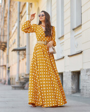 Pretty Stylish Woman Wearing Yellow And White Polka-dot Sundress And Sunglasses. Beautiful Girl In Long Dress Walking And Standing At City Street