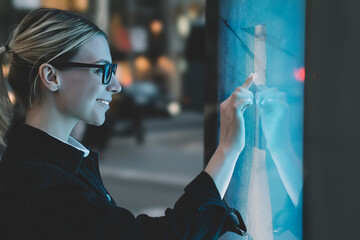 Wall Mural - Smiling female standing at big display with advanced digital technology. Young woman touching with finger sensitive screen of interactive kiosk for find information while standing on street in evening