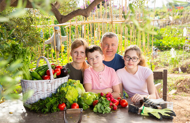 Portrait of happy family with two kids posing in garden with fresh harvest of vegetables and greens