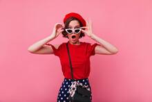 Surprised French Girl In Stylish Sunglasses Posing On Pink Background. Indoor Photo Of Elegant White Woman In Red Clothes.