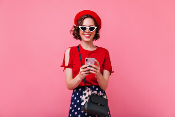 Excited european woman in elegant french outfit texting message. Adorable caucasian girl in sunglasses and beret posing with phone.