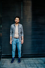 Full length portrait of caucasian hipster guy dressed in stylish casual wear standing against promotional background and looking at camera.Young bearded man spending leisure time outdoors