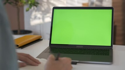 Young student looking at green screen laptop computer in living room watching education video content online. Over shoulder close up view. Business woman or female freelancer working from home office.