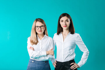 The theme is a female team. Portrait of two young caucasian business women posing in studio on a colored background. Teamwork two people in office clothes on a turquoise wall