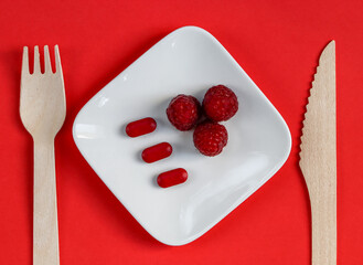 strawberry on a plate.
Three red pills and three raspberries in a white saucer on a red background, top view close-up.