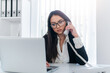 © Bojan Topalovic - Busy lady in suit with glasses talking on the phone and looking at laptop on the desk.