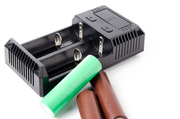 A black charger for two 18650 batteries. A green and brown 18650 battery on a white background next to the discharge, on charging. Male hand puts the battery on charge