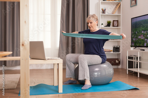 60 years old woman exercising from home. Old person pensioner online internet exercise training at home sport activity with dumbbell, resistance band, swiss ball at elderly retirement age