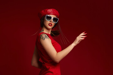 Wall Mural - stylish brunette woman in white sunglasses, hat and dress posing in studio on red background. woman looking back