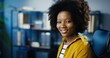 © VAKSMANV - Close up portrait of pretty African American girl smiling to camera while sitting at office indoors. Beautiful cheerful curly young woman in good mood with smile on face in cabinet. Emotions concept